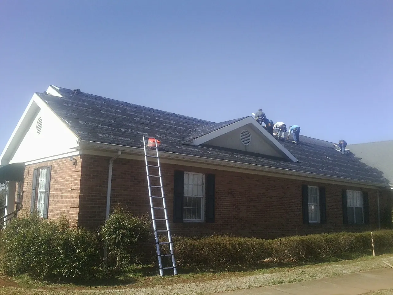 Crew tearing shingles off a brick ranch roof with a ladder set — Metal Roof Repair work by our crew in Elbert
